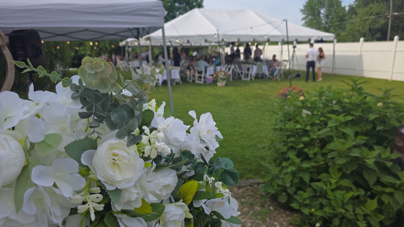 Guests at an outdoor party under white tents on a lawn, with white flowers in front