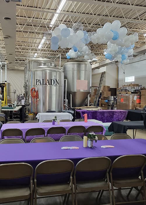 Purple-covered banquet tables set up in front of the Paladin brewing tanks, with balloon garlands overhead